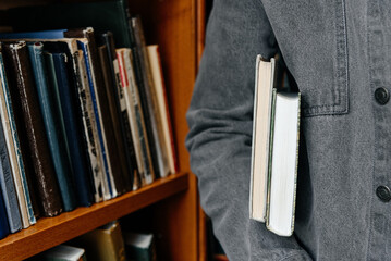young man in grey jacket holds stack of books ander arm, bookshelves in background, student...