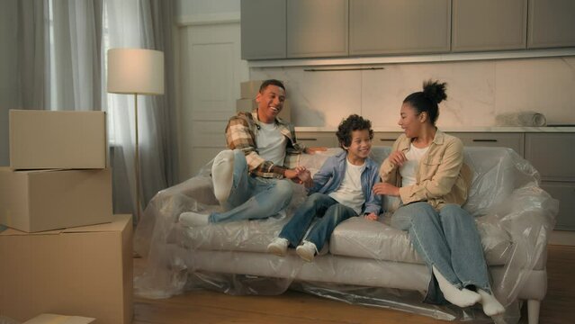 Happy carefree African American family home owners mom dad little boy kid child son jumping on sofa couch together moving in new house celebrate relocation renovation buying flat rent mortgage credit