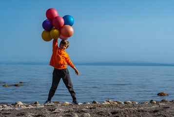 Cheerful boy standing with balloons on lake shore on summer day