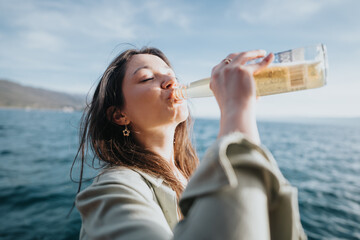 A happy girl blissfully drinks a cold beer, savoring the moment by the ocean under a clear sky.