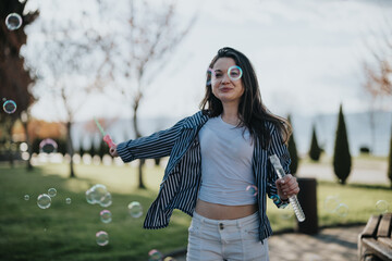 A joyful young woman in a striped jacket and white pants playing with soap bubbles in a park on a...