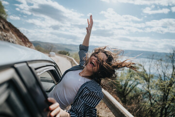 Joyful young woman with wild hair streaming, expressing freedom while leaning out of a car on a mountainous road trip.