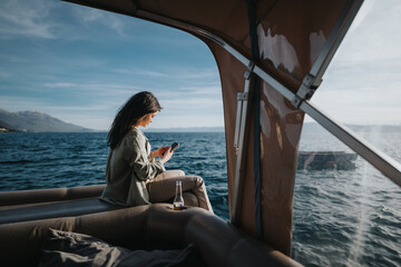 Happy young woman enjoys a boat ride, using her phone with a scenic lake background and clear blue...