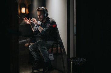 Man with headphones gesturing while gaming at night, displaying emotion and focus, in a moody lit...