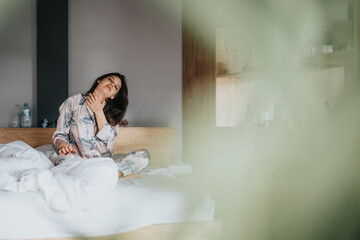 A young woman wearing pajamas is captured mid-stretch in a cozy bedroom, expressing relaxation and...