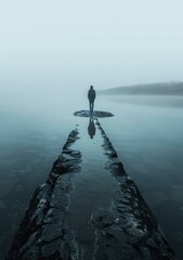 b'Man standing alone on a pier surrounded by water and fog'