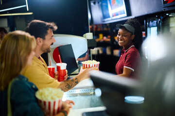 Happy black concession stand worker selling drinks and popcorn before movie projection in cinema.