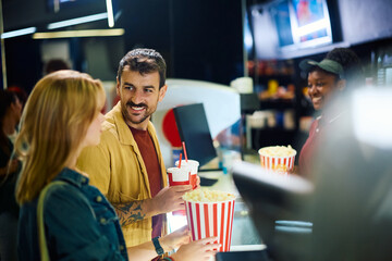 Smiling man and his girlfriend buying drinks and popcorn in cinema.
