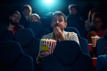 Young man watching suspenseful film projection in movie theater.