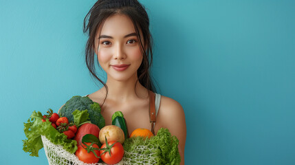 Indoor shot of cheerful dark haired Asian woman holds freshly made smoothie from vegetables carries net bag wears cropped top and leggings isolated over blue background blank space for promo. - obrazy, fototapety, plakaty