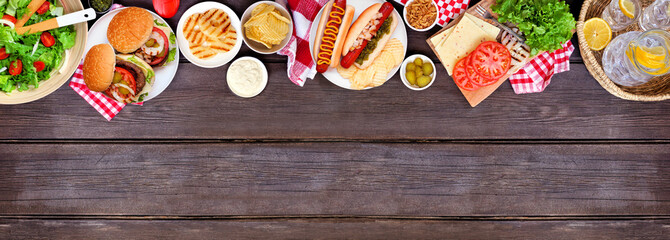 Summer BBQ or picnic top border with hamburgers, hotdogs, salad and snacks. Above view over a dark wood banner background. Copy space.