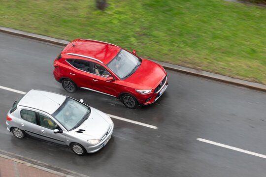 Red Chinese MG ZS crossover overtaking Peugeot 206 car in heavy rain on street