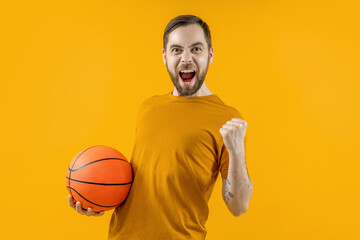 Studio portrait of young attractive basketball player or supporter posing over bright colored orange yellow background holding the ball in hand and making winner's gesture clenching his fist