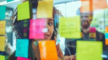 Curly-haired woman placing colorful sticky notes on glass partition. Bright office setting with male colleague in the background.