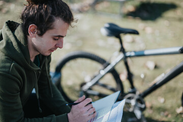 A serene scene of a young man relaxing by his bike in a peaceful park, thoughtfully writing or...