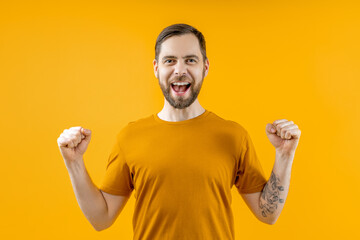 Studio portrait of bearded attractive joyful young man clenching his fists in winners gesture while posing over yellow background