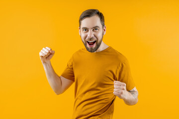 Studio shot of bearded attractive joyful young man clenching his fists in winners gesture while posing over bright colored orange yellow background