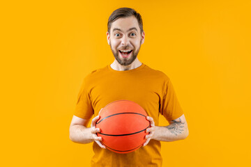 Studio portrait of young attractive excited basketball player or supporter posing over bright colored orange yellow background with the ball in hands