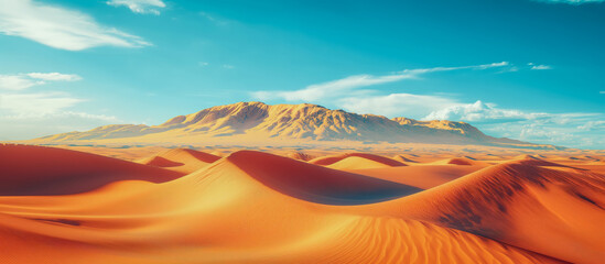 Breathtaking landscape of rolling orange sand dunes under a clear blue sky with a rugged mountain range in the background, capturing the vastness of the desert
