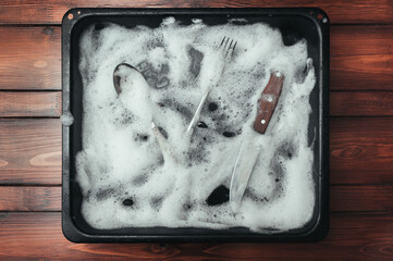 A wooden knife, fork and spoons in the detergent foam on a black oven-tray. Washing dishes concept.