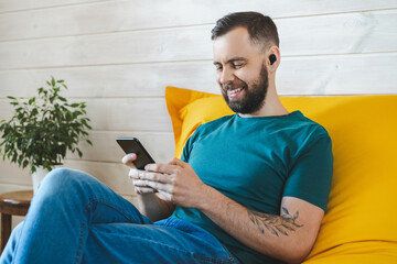 Portrait of smiling bearded man in wireless earphones is listening to music and surfing internet on his mobile phone while sitting at home on comfortable yellow sofa