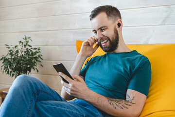 Portrait of young handsome casually dressed smiling man having video call via his mobile phoner while sitting on comfortable yellow sofa at home