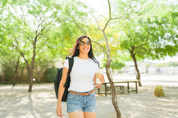 Happy young hispanic woman in a white t-shirt and sunglasses, walking in a sunny park with a backpack and coffee