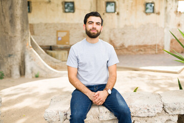 Young hispanic man in a simple grey t-shirt sitting outdoors with a casual smile, ideal for showcasing apparel mockups