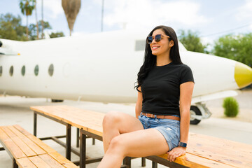 Confident latin woman wearing a black t-shirt and sunglasses sits casually in front of an airplane on a sunny day