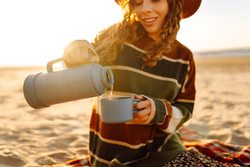 Happy woman in a stylish sweater sits at picnic on the beach drinks a hot drink from a thermos and...