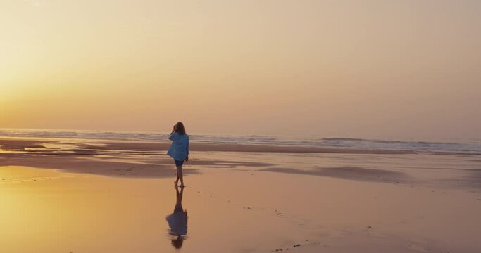Cinematic shot of serenity and tranquility as young woman walks towards setting sun on empty beach during low tide. Golden hour at amazing location. Peaceful connection with yourself and nature