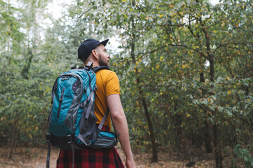 Young man with tourist backpack walking alone in early autumn forest and looking to the side