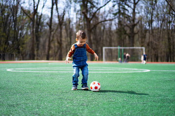 A young boy in overalls is playing with a soccer ball on a green field. The boy is wearing a red shirt and blue jeans