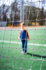 A young child stands in front of a soccer goal. The child is wearing a blue overalls and a red shirt.