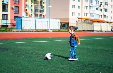 A young boy is playing with a soccer ball on a field. The boy is wearing a blue overalls and is...