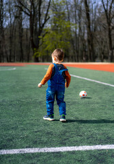 A young boy stands on a soccer field with a soccer ball and a second ball. The boy is wearing a blue overalls and an orange shirt