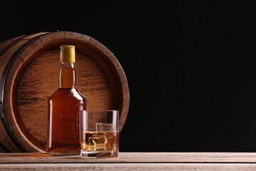 Whiskey with ice cubes in glass, bottle and barrel on wooden table against black background, space...