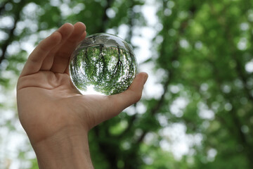 Beautiful green trees outdoors, overturned reflection. Man holding crystal ball in park, closeup....