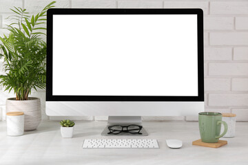 Office workplace with computer, cup, glasses, stationery and houseplant on light table near white brick wall