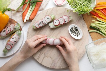 Woman wrapping spring roll at white wooden table with products, top view