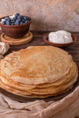 A plate of golden pancakes topped with blueberries, accompanied by a bowl of sour cream, a portion of fresh cottage cheese and a glass of milk. On a beige-golden background with a green runner.