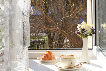 Cup of tea,croissant and spring flowers in a vase on a sunny window.The concept of home comfort and greeting of spring.Beautiful flower arrangement with hyacinths,selective focus.