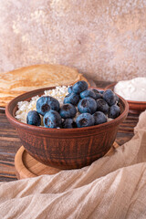 A plate of golden pancakes topped with blueberries, accompanied by a bowl of sour cream, a portion of fresh cottage cheese and a glass of milk. On a beige-golden background with a green runner.