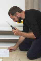 Man on the floor assembling furniture at home, holding a screwdriver and looking at assembly instructions. There is a partially assembled wooden shelves, a yellow level tool nearby. Screwdriver work