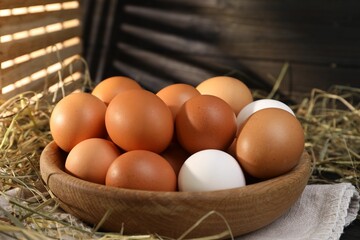 Fresh chicken eggs in bowl and dried hay on table, closeup