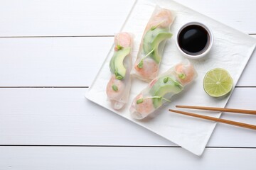 Delicious spring rolls, lime, soy sauce and chopsticks on white wooden table, top view. Space for...