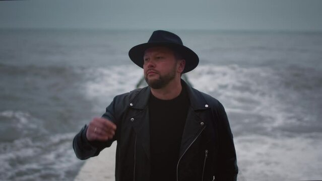 Stylish man in a leather jacket and hat stands on a breakwater. Male walks along the seawall, strong and big waves on the sea or ocean. Cloudy weather in winter or autumn. Human artist in a rock band