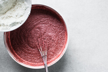 Top view of flour being folded into Red velvet cake batter in white ceramic mixing bowl, process of...