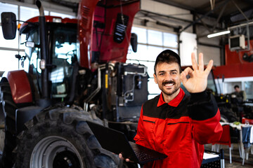 Experienced mechanic with diagnostic tool standing by tractor machine inside workshop.
