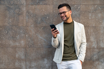 Happy handsome man using mobile phone while standing in front of a wall, sending a text message or...